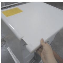 Hand holding a white plastic tray with a yellow sticker, partially visible in a workshop setting (no text or symbols on the tray itself)