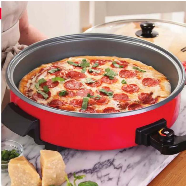 Red pizza cooker with cheese and green herbs, placed on a patterned cloth (no text or symbols visible)