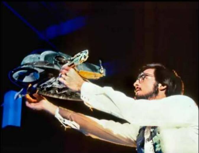 Man in white shirt and glasses handling a large metallic object on stage (no visible text or symbols)