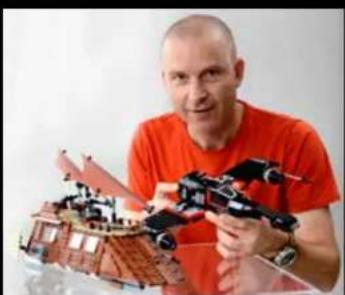 Man in orange shirt holding a model of a drone-like device with a red roof and black components (no visible text or symbols)