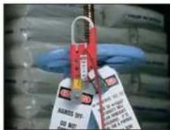 Close-up photo of a hand holding a red and white safety tag with visible text, partially obscured by blue gloves.