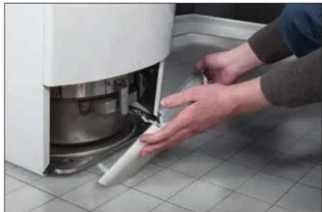 Person using a tool to clean or install a white washing machine component on tiled floor (no text or symbols visible)