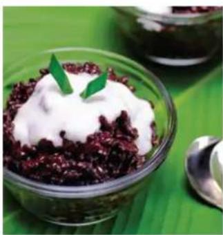 Close-up of a glass bowl containing a dark red rice dessert topped with white whipped cream and a green garnish, placed on a green surface with a spoon nearby (no text or symbols visible)