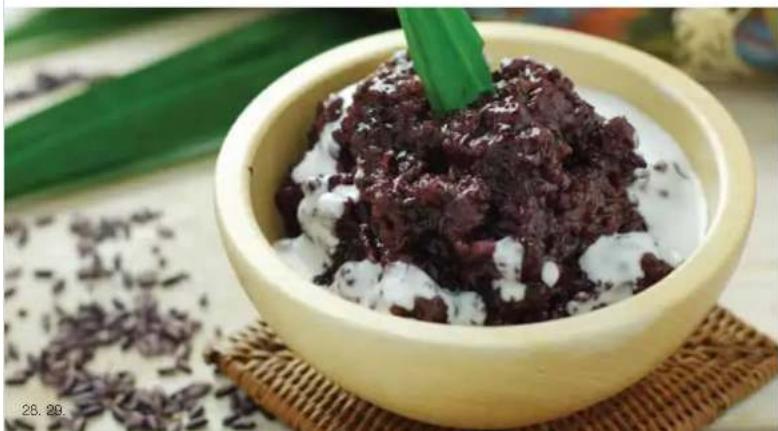 Bowl of dark brown rice topped with white grains, placed on a woven mat with scattered dried herbs in the background (no text or symbols visible)