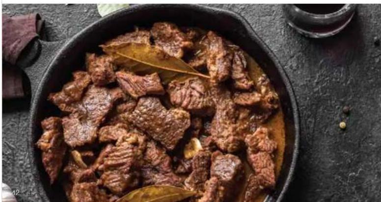 Close-up of a braised meat dish in a black pot with visible herbs and spices, placed on a dark textured surface (no text or symbols)