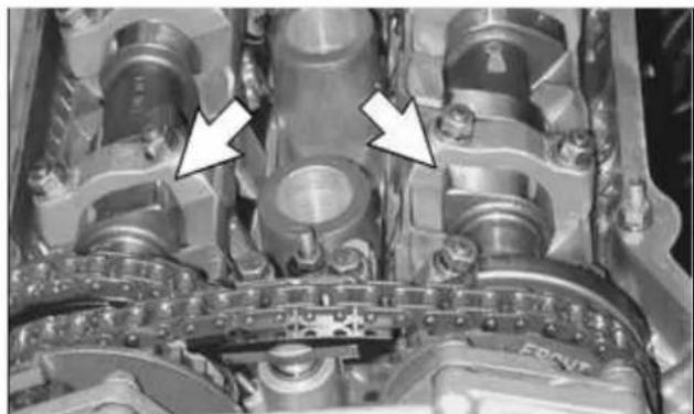 Close-up of an internal combustion engine cylinder head with two white arrows pointing to specific components (no text or symbols visible)