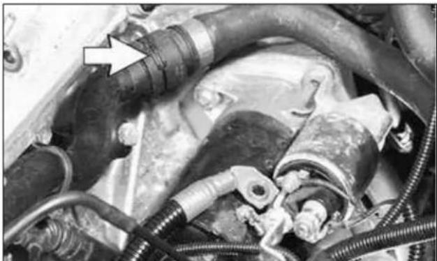 Close-up of automotive engine components with hoses and a white arrow pointing to a component (no visible text or symbols)