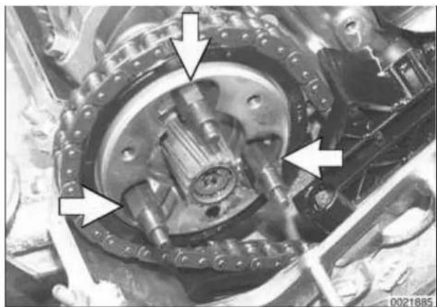 Close-up mechanical assembly showing a central gear-like component with bolted joints and mounting flanges, marked with white arrows (no visible text or symbols)