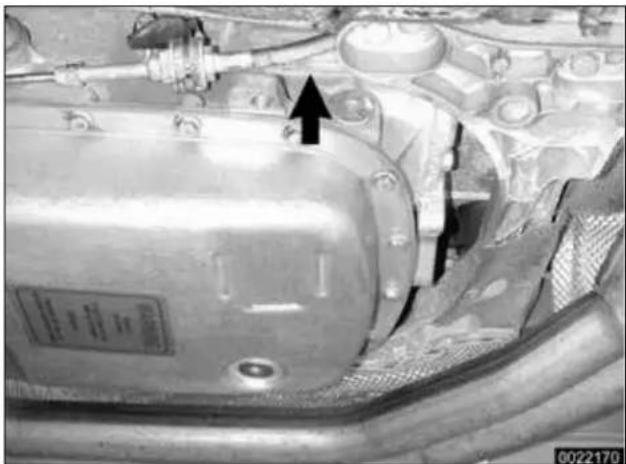 Close-up of a metallic automotive engine component with mounting flanges and a black arrow pointing to a component (no visible text or symbols)