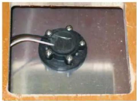 Close-up of a black circular mechanical component with four bolts, mounted on a dark rectangular base (no visible text or symbols)