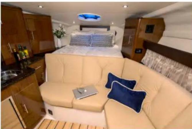 Interior view of a modern boat cabin featuring a beige sofa, cream-colored bedding, and a blue cushion with a laptop (no visible text or symbols)