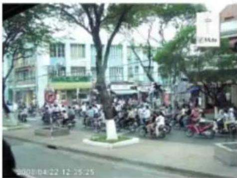 Street photo showing a crowded urban street with people on motorcycles and trees, featuring visible building signage and timestamp at bottom