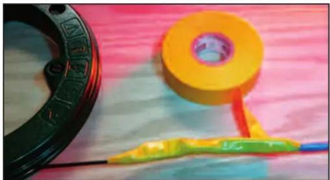 Close-up of a yellow tape measure tool with a red ring and green handle, next to a black circular object on a textured surface (no text or symbols visible)
