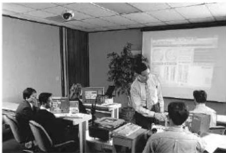 Black-and-white photo of an office meeting or presentation with a presenter and projection screen showing charts and data