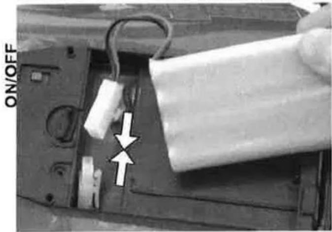 Close-up of a mechanical component with a white cylindrical body and a curved hook, showing a white arrow pointing to a specific part (no text or symbols visible)