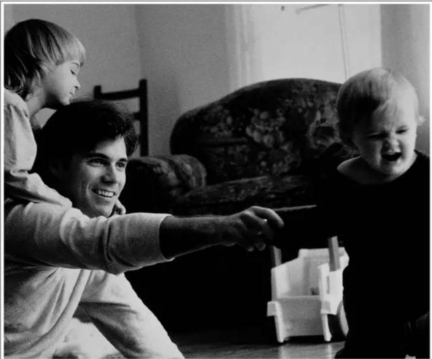 Black-and-white photo of a man and two children playing together, one pointing at a small model on the other (no text or symbols visible)