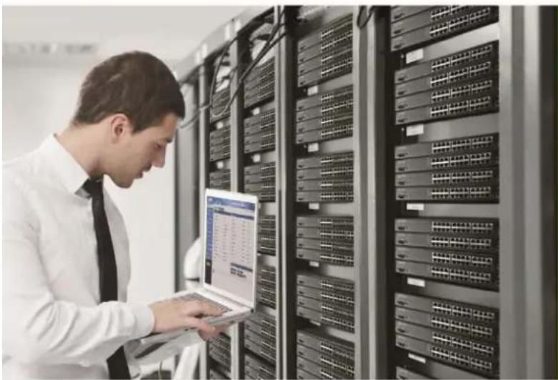 Man in white shirt using laptop in server rack with network switches (no visible text or symbols)