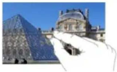 Hand holding a glass pyramid next to the Louvre Pyramid building under a clear blue sky (no text or symbols visible)