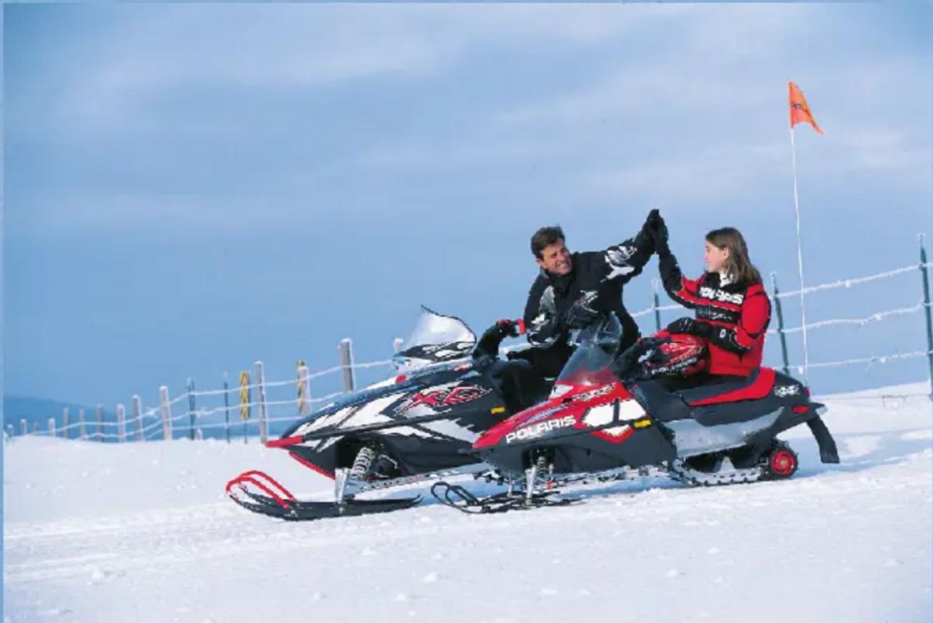 Two people riding SnowPARIS scooters on a snowy slope under clear blue sky, with barbed wire fence and distant hills in background (no text or symbols on main subjects)