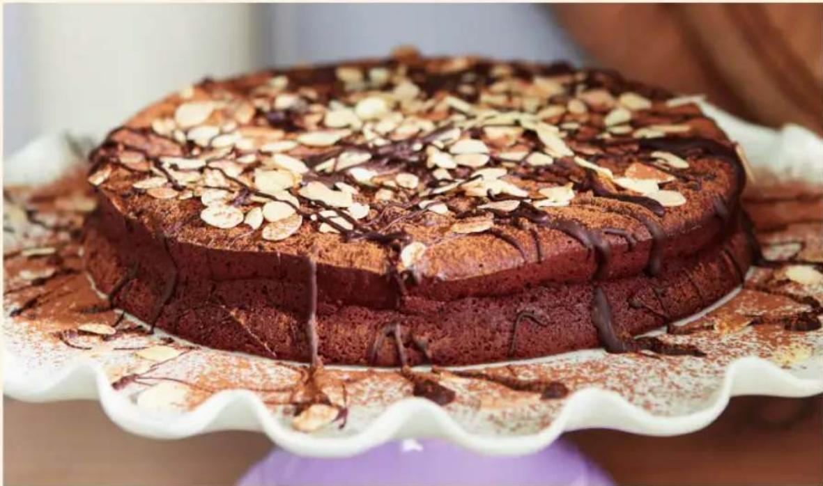 Close-up of a chocolate cake with almond slices and chocolate drizzle on a white scalloped plate (no text or symbols visible)