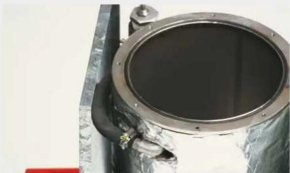Close-up of a metallic cylindrical mechanical component with a black central opening, attached to a metal frame (no visible text or symbols)