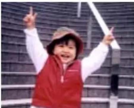 A smiling child in a red vest and hat, posing with raised fists in front of a brick wall (no text visible)