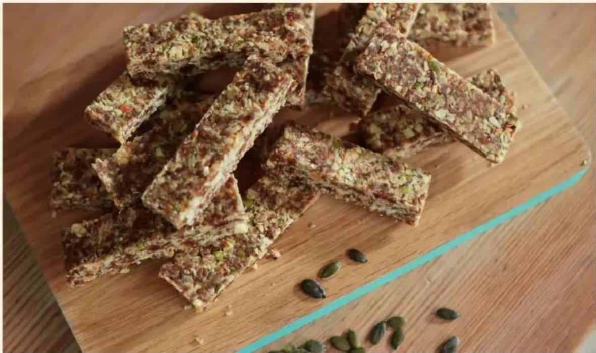 Stacked rectangular food items on a wooden cutting board, garnished with chopped green onions and nuts, with scattered seeds nearby (no text or symbols visible)