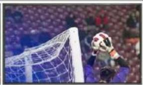 Soccer goalkeeper standing at the goal with a soccer ball in the air, against a blurred stadium background (no visible text or symbols)