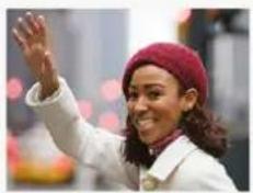 Woman wearing a red beanie and white coat waving, cityscape background (no visible text or symbols)