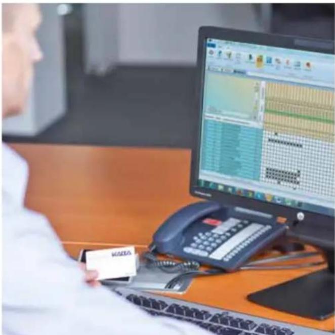 Office scene with person using a smartphone displaying a financial chart on a computer screen, showing data tables and interface elements.