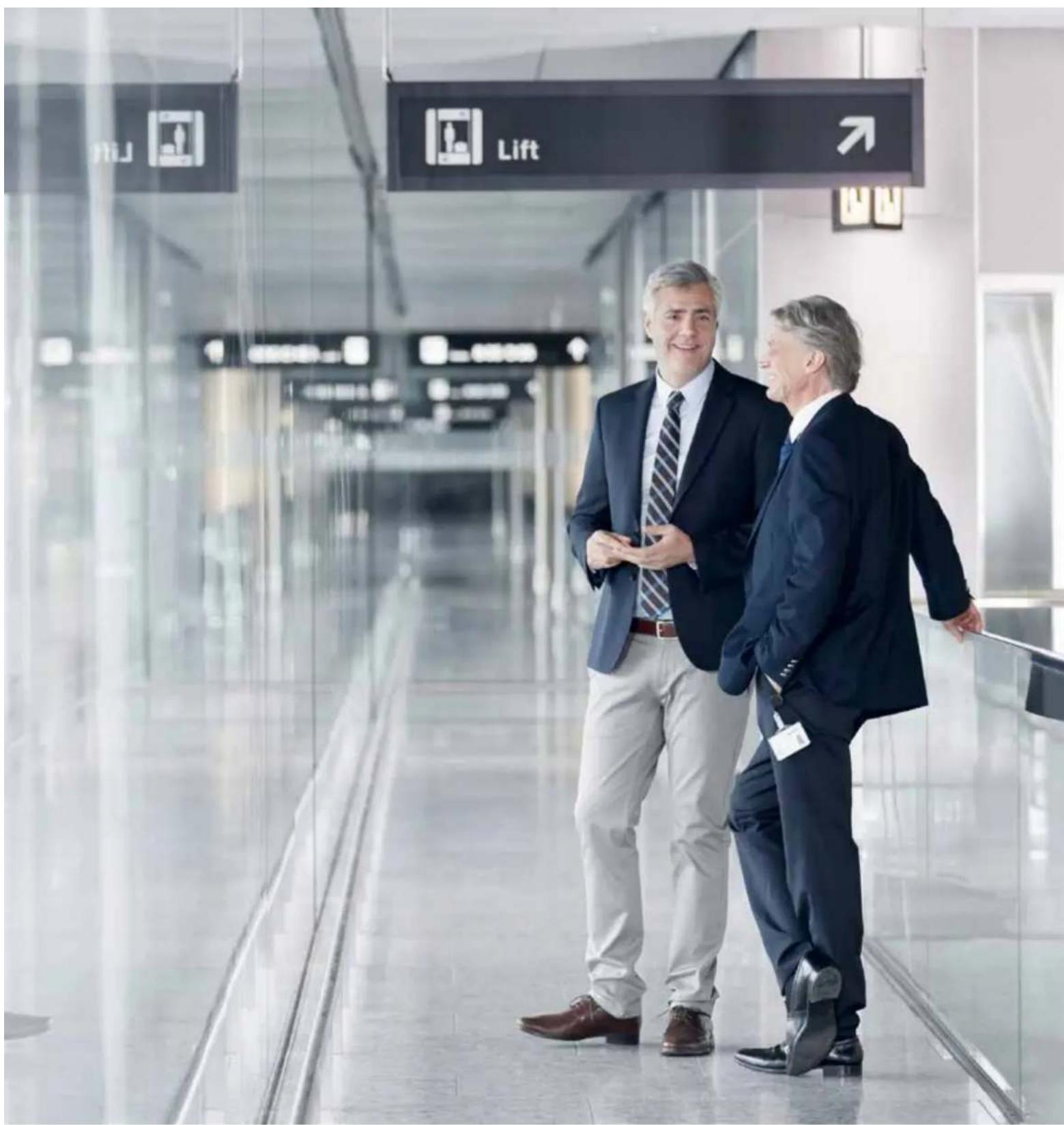 Two men in suits walking on a modern corridor, with visible directional signs including 'Lift' and 'HJ', indicating transportation or transit directions.