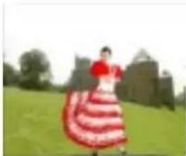 Person in red and white striped dress dancing outdoors on a grassy field with trees and buildings in the background (no visible text or symbols)