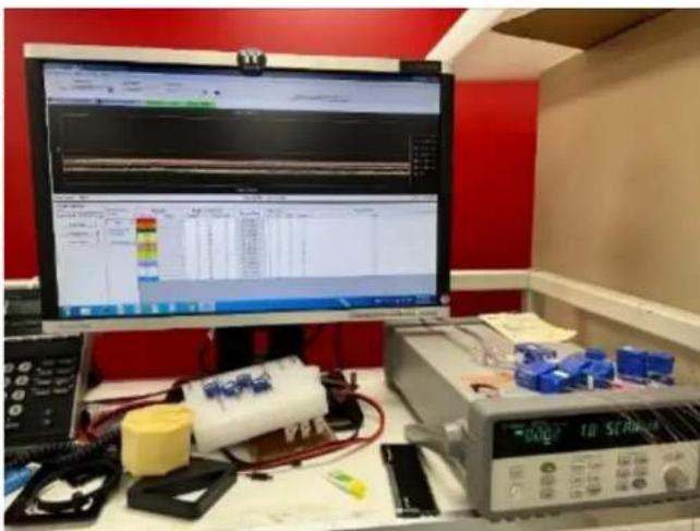 Lab setup with computer monitor displaying a technical schematic, surrounded by electronic devices and tools including a battery and oscilloscope.