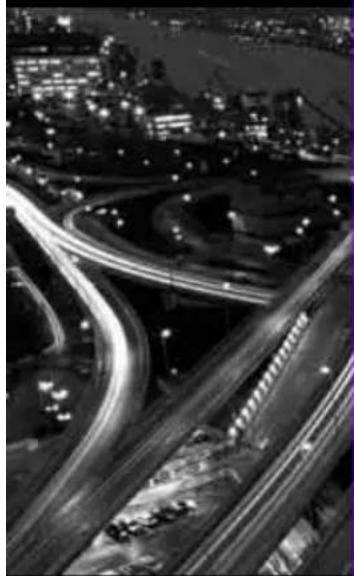 Nighttime aerial view of a complex highway interchange with illuminated light trails and surrounding urban buildings (no visible text or signage)