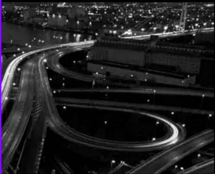 Nighttime aerial view of a complex highway interchange with illuminated light trails, surrounded by city lights and water (no visible text or signage)