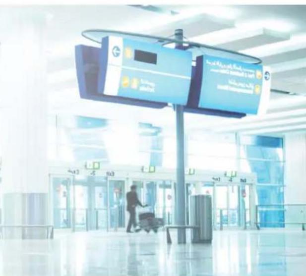 Airport terminal interior with digital kiosk displaying 'Kuwaiti Bank' signage and a person pushing a cart in the distance.