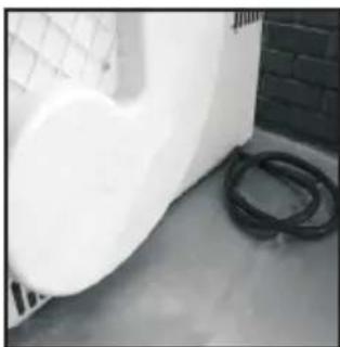 Exterior view of a white industrial or laboratory equipment with a coiled black pipe, next to a grid-patterned wall (no visible text or symbols)