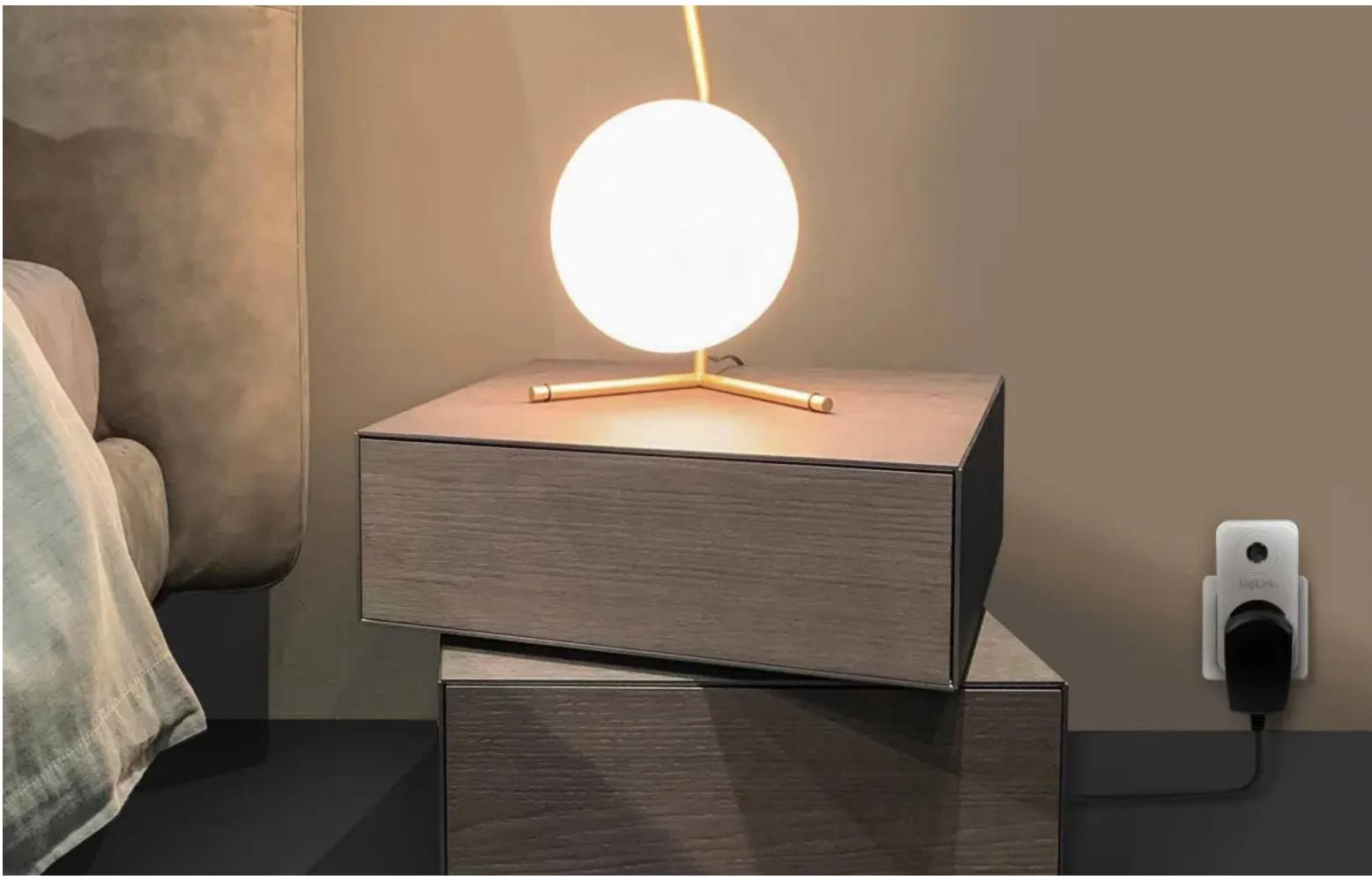 Modern minimalist desk lamp with a glowing circular light source, placed on a matching wooden side table next to a bed and a wall-mounted power outlet (no text or symbols visible)