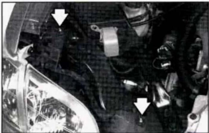 Close-up of a car engine bay with visible structural components and two white arrows pointing to specific parts (no text or symbols present)