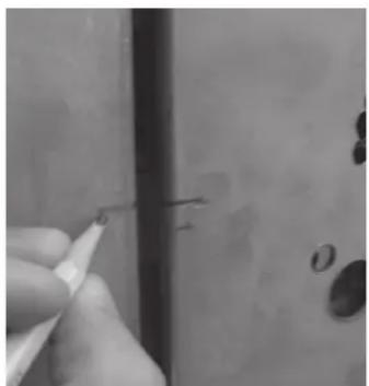 Close-up of a hand holding a pen, pointing at a small object on a wooden surface (no visible text or symbols)