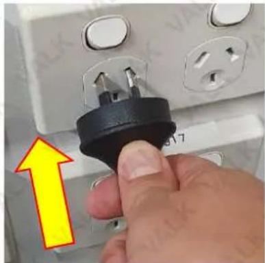 Close-up of a hand inserting a black plastic plug into an electrical outlet socket, with a yellow arrow pointing to the plug (no text or symbols visible)