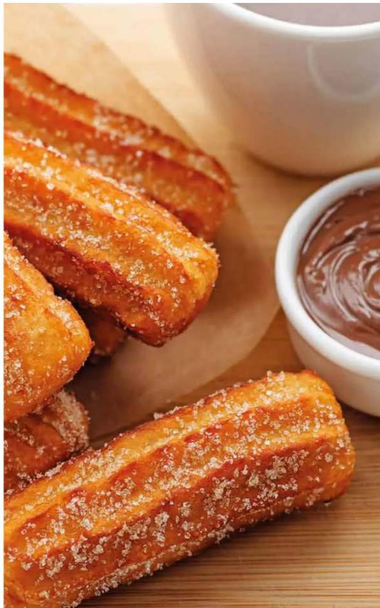 Close-up of golden-brown fried pastries with powdered sugar, served on a wooden board next to a small bowl of chocolate sauce (no text or symbols visible).