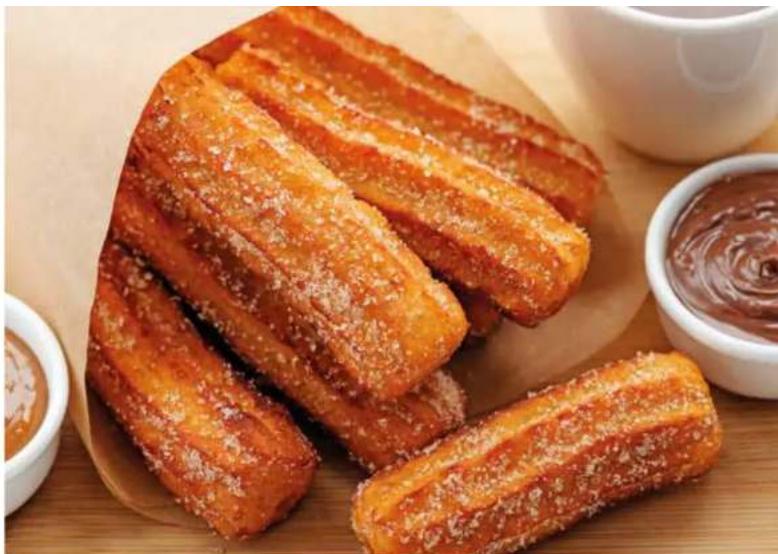 Close-up of golden-brown fried pastries with powdered sugar, served on a wooden board with small bowls of sauce in the background.