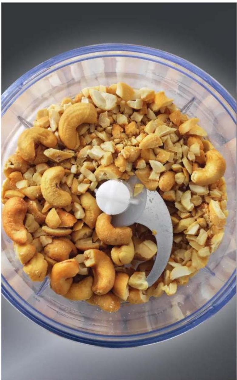 Top-down view of a wooden mixer bowl containing a pile of cashew nuts and nuts, with a white plastic tool partially visible (no text or symbols)