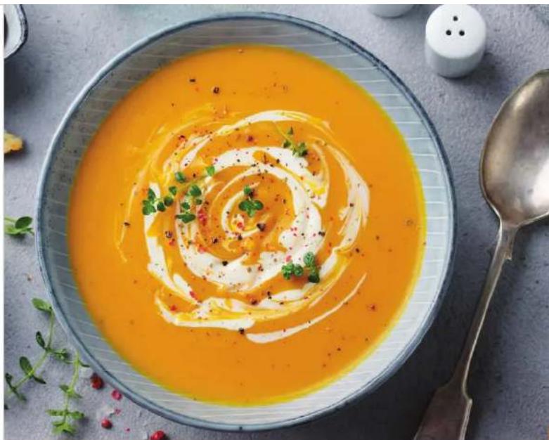 Bowl of creamy orange soup with spiral swirl garnish, served on a metal plate with a spoon and small figurines nearby (no text or symbols visible)