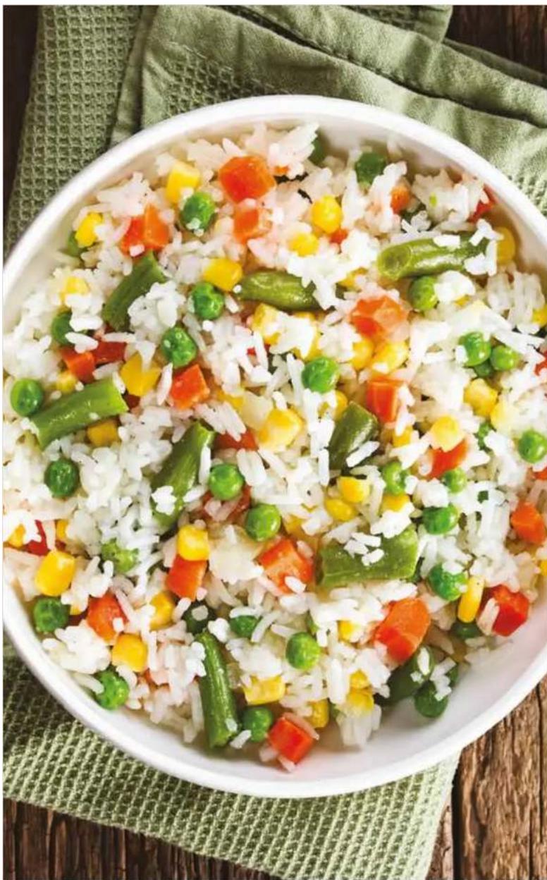 A bowl of stir-fried rice with vegetables and carrots, placed on a woven mat with a green cloth background (no text or symbols visible).