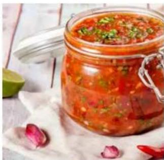 Glass jar filled with red sauce and chopped green herbs, placed on a white cloth with scattered red rose petals (no text or symbols visible)