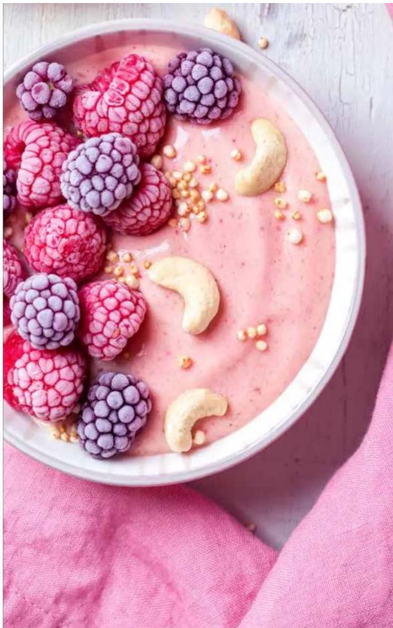 Bowl of pink smoothie with purple berries and cashew nuts, placed on a pink cloth (no text or symbols visible)