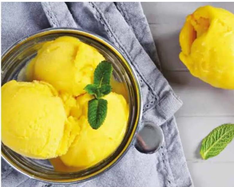Close-up of yellow ice cream scoops in a metal bowl with mint leaves, placed on a cloth with a spoon and leaf nearby (no text or symbols visible)