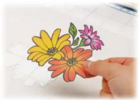 Hand holding a colorful paper craft of daisies (yellow, orange, pink) on grid paper, no text or symbols visible